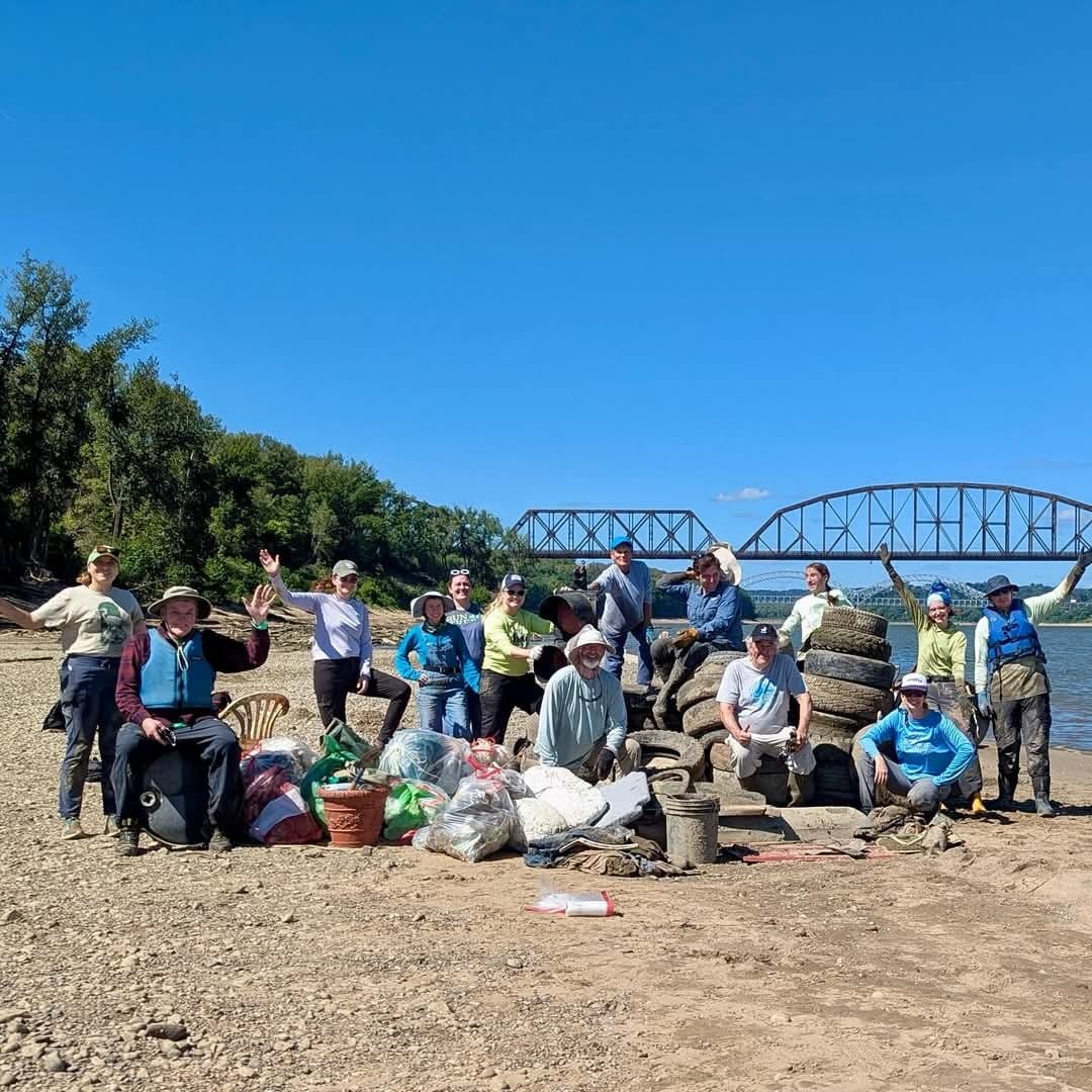 A group of people pose on a rocky riverbank with collected trash, including tires and bags, after a cleanup effort. A steel bridge and trees are visible in the background under a clear blue sky.