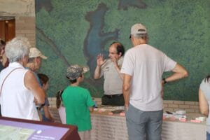 A park ranger speaks to a group of adults and children gathered around a table with display items, in front of a large map or nature mural.