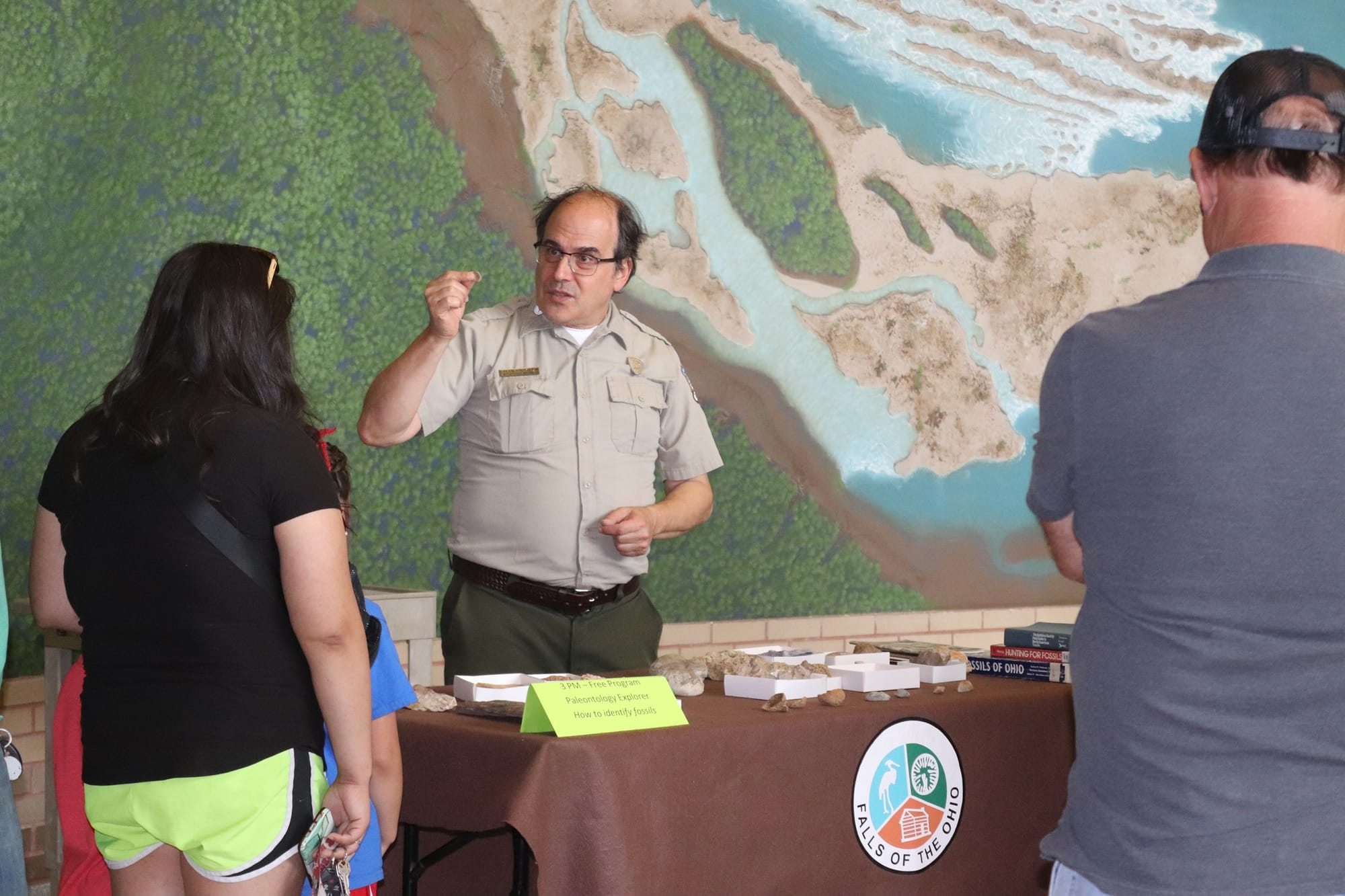 Alan Goldstein fossil ID best 2000 A park ranger speaks to a group of people at an indoor educational display table with rocks and books, in front of a large map or diorama of a river and land. A yellow sign sits on the table.