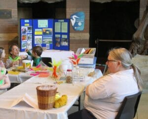 A group of people sit at a table covered with craft supplies, feathers, and baskets. A display board with information and photos stands behind them. They appear to be participating in an indoor activity or workshop.