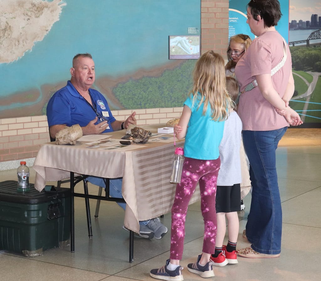Dale Brown Six Fossils program 3-30-24 1024 A man sits at a table with fossils, talking to a woman and three children standing in front of him at a museum or educational event. The group appears engaged in conversation.