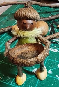 A small figure made from acorns, twigs, and nuts stands on a green cutting mat, holding a large acorn cap with twig arms. The figure has a fluffy yellow collar and nut-like shoes. Craft supplies are visible in the background.