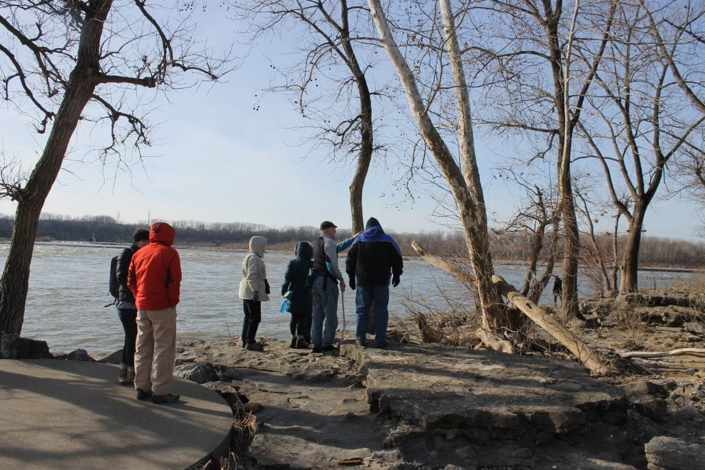 A group of people dressed in winter clothing stand on a rocky riverbank surrounded by leafless trees, looking out over the water under a clear sky.
