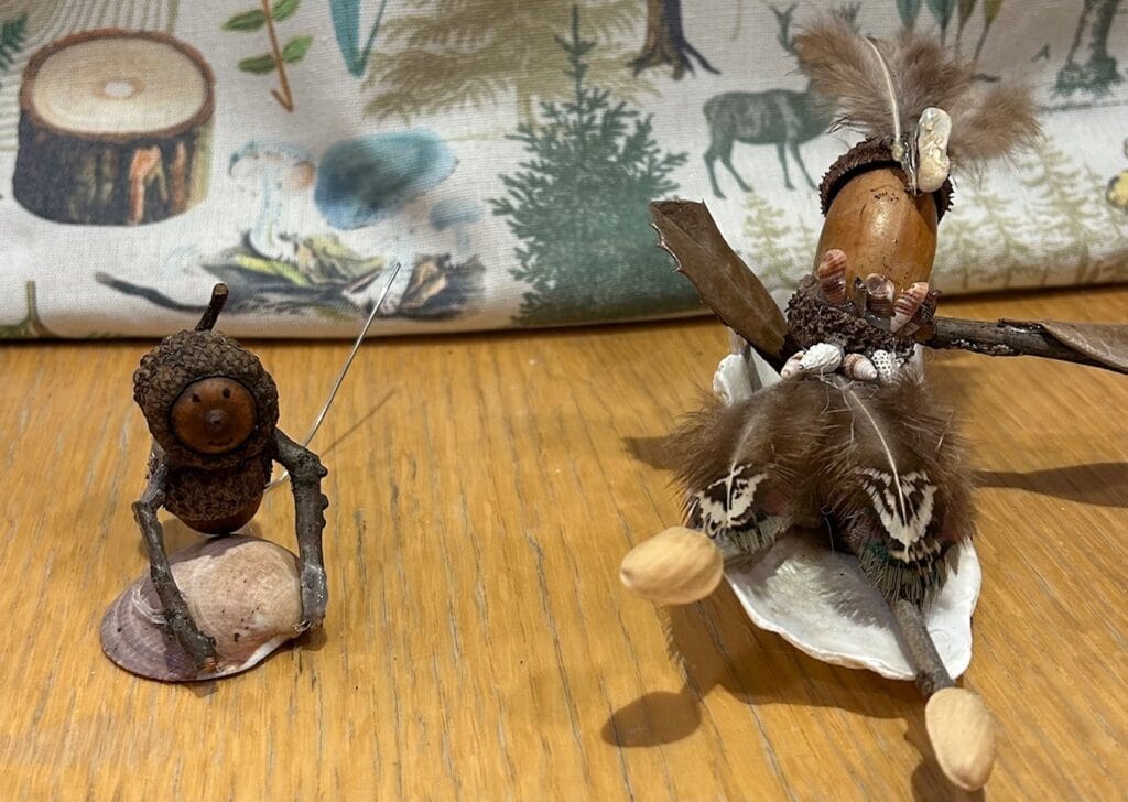 Two small handmade figurines crafted from natural materials like acorns, shells, feathers, and sticks, are placed on a wooden surface with a forest-themed fabric in the background.