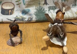 Two small handmade figurines crafted from natural materials like acorns, shells, feathers, and sticks, are placed on a wooden surface with a forest-themed fabric in the background.