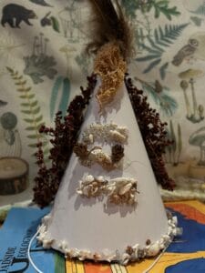 A handmade white cone decorated with tiny shells, nuts, and seeds, with dried red berries along the sides and a feather on top, placed against a nature-themed background.