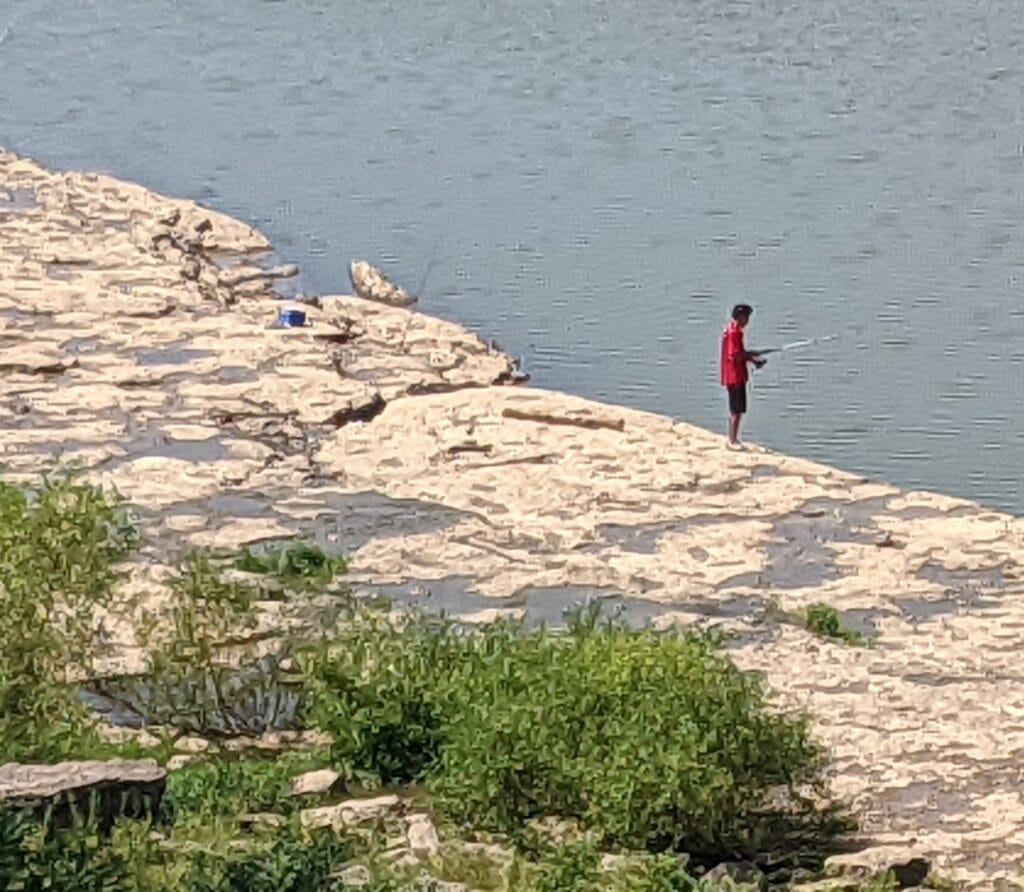A person wearing a red shirt stands on rocky ground by a river, fishing with a rod. Green bushes are visible in the foreground.