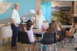 Two adults wearing name tags stand by a table, speaking to a group of children who are seated and raising their hands, in a classroom with maps and photos on the walls.
