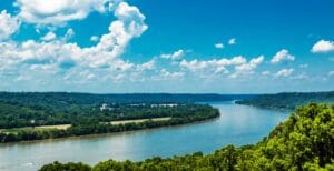 A wide river winds through lush green forests under a bright blue sky with scattered clouds, creating a peaceful and scenic landscape.