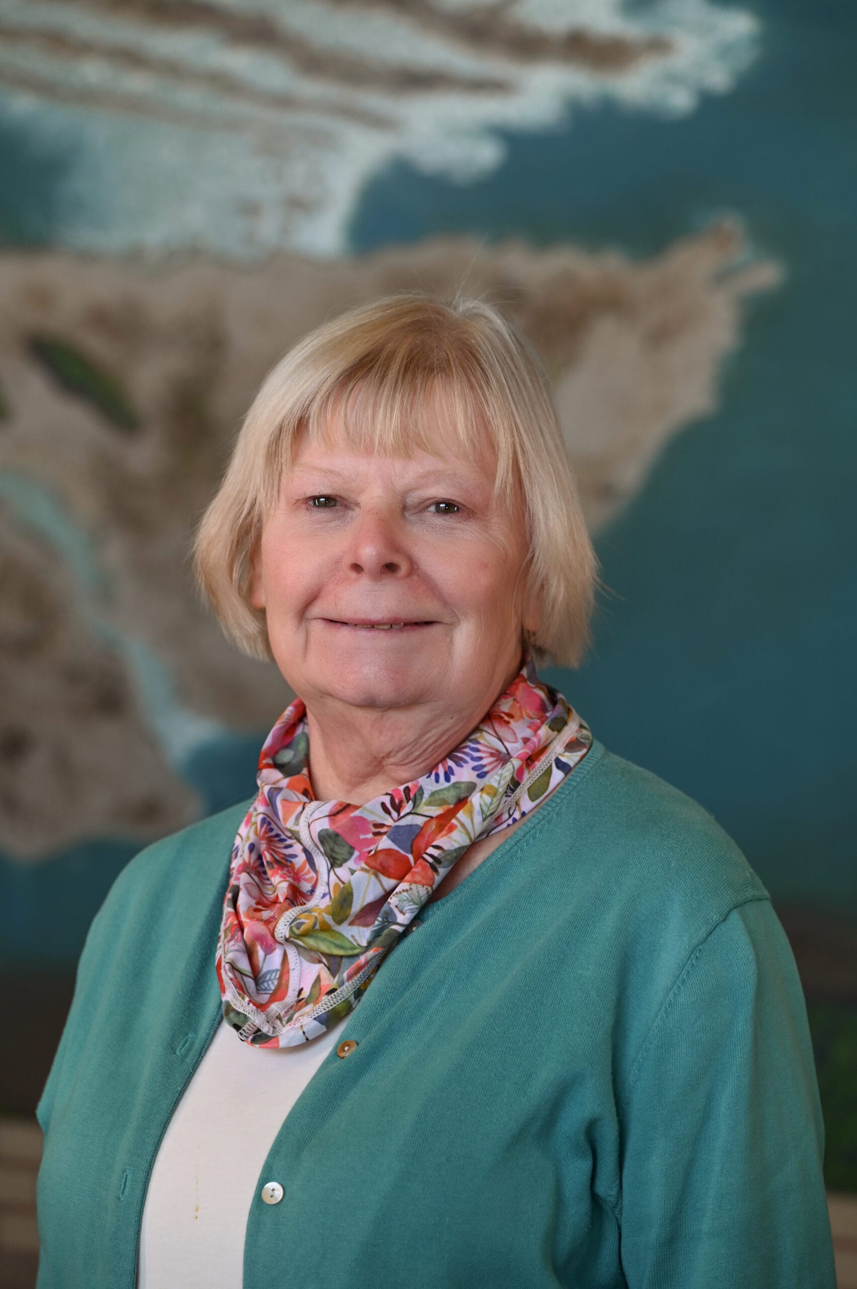 An older woman with short blonde hair, wearing a teal cardigan and a colorful floral scarf, smiles in front of a blurred map background.