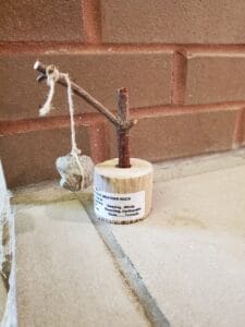 A small weather rock display with a stone tied to a string, hanging from a twig mounted on a wooden base. A humorous label explains how to interpret the rock’s movement as weather conditions. Brick wall in the background.
