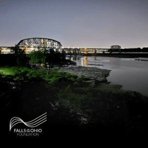 A nighttime view of a bridge with lights reflecting on the river below, surrounded by trees and dark greenery. The “Falls of Ohio Foundation” logo appears in the lower left corner.