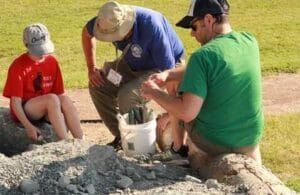 Three people, two adults and a child, examine rocks and fossils outdoors. They are sitting and crouching near a pile of gravel, using tools and a white bucket, on a sunny day with grass in the background.