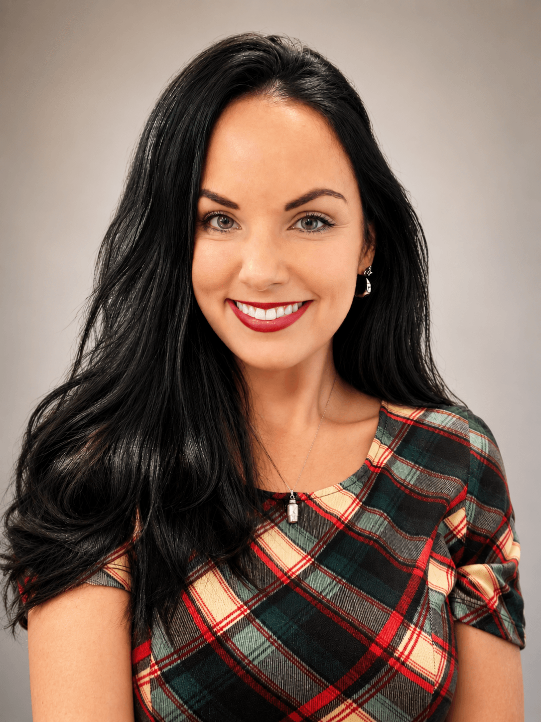 A woman with long black hair and green eyes smiles at the camera. She is wearing a plaid dress in green, yellow, and red colors, along with a necklace and earrings. The background is plain and softly lit.