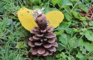 A craft figure made from a pinecone body, acorn head with drawn face, yellow leaf wings, and a small flower crown sits on green grass and plants outdoors.