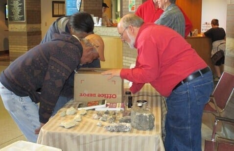 Three men lean over a table covered with rocks and minerals, closely examining the items. One man in a red shirt gestures toward the display, which is set up indoors with a cardboard box and magnifying glass on the table.