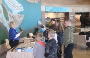 A woman shows an artifact to a group of children standing in front of her in a museum setting, with a large map on the wall and tables displaying various objects.