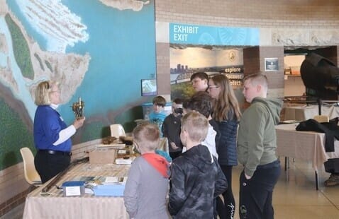 A woman shows an artifact to a group of children standing in front of her in a museum setting, with a large map on the wall and tables displaying various objects.