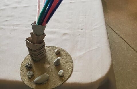 A craft palm tree made from stacked cardboard and colored pipe cleaners stands on a sand-colored circular base decorated with small stones.