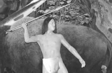 A black and white image of a mannequin dressed as a prehistoric human holding a spear, posed in front of a model of a large animal, possibly for a museum display.