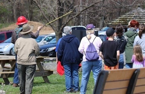 A group of people, seen from behind, watch a person in a red helmet pointing with a long stick outdoors near parked cars and trees.