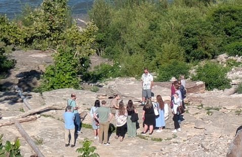 A group of people stand on a rocky area surrounded by greenery, listening to a guide. The guide is standing in the center, while the group forms a loose circle around him. The scene is outdoors near a river.
