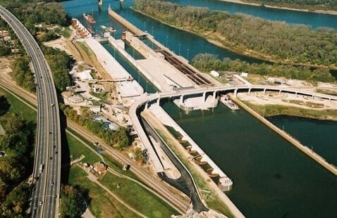 Aerial view of a river lock and dam system, with roads, water channels, and green areas nearby. Multiple lock chambers and gates are visible, along with a bridge crossing over the site.