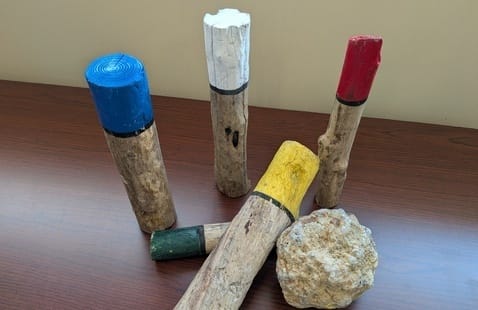 Five pieces of wood painted blue, white, red, green, and yellow on one end, standing or lying on a wooden surface next to a rough rock.