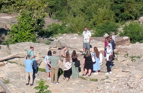 A group of people standing on a rocky outcrop outdoors, listening to a man speaking.