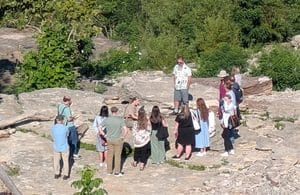 A group of people standing on a rocky outcrop in a wooded area, listening to a man in a patterned shirt.