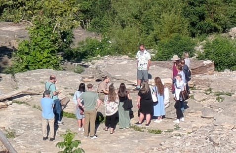 A group of people standing on a rocky outcrop in a wooded area, listening to a man in a patterned shirt.
